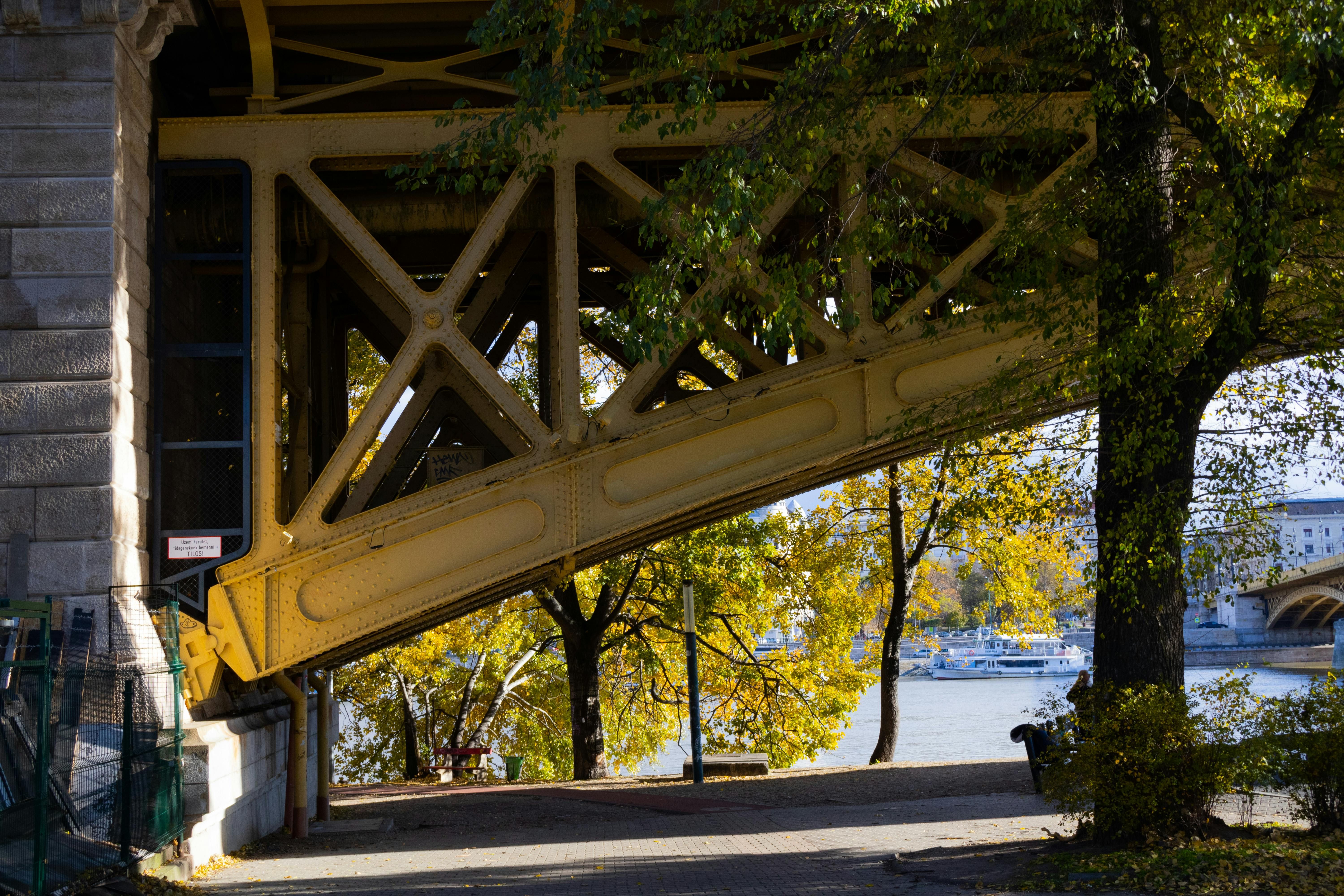 Yellow metal bridge structure extends over a path, with autumn trees and a body of water.